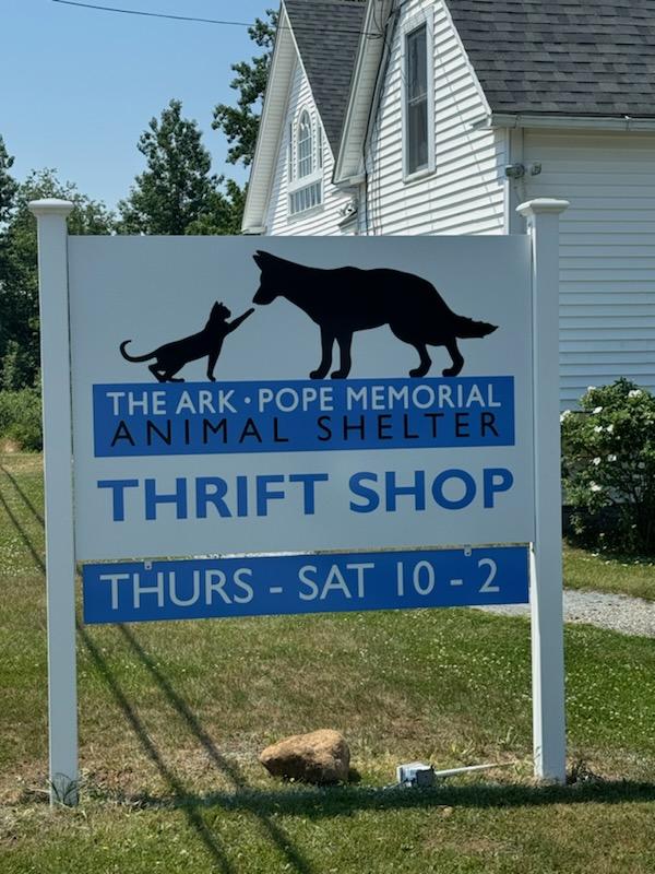 The Ark Pope Memorial Animal Shelter Thrift Shop sign displaying the shop's name and hours: Thursday to Saturday, 10 AM to 2 PM, featuring a black cat and dog silhouette on a white background.